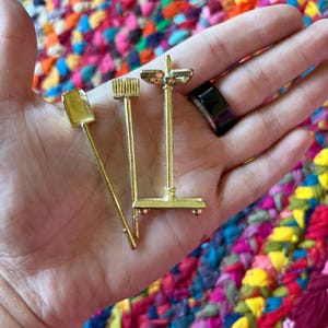 Peut inclure: Un ensemble de trois outils de cheminée miniatures de couleur dorée comprenant une pelle, une brosse et un support. Les outils sont métalliques avec une finition lisse et sont conçus comme des accessoires à petite échelle pour une maison de poupée.