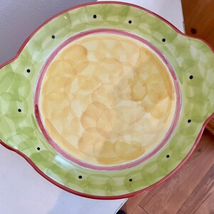May include: A large, round serving platter with a scalloped edge. The platter is decorated with a green and yellow pattern and has a red rim. There are black dots around the edge of the platter.