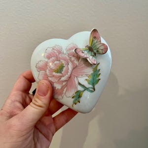 Puede incluir: Una caja de porcelana blanca en forma de corazón con un diseño floral rosa y verde y una mariposa en la parte superior.