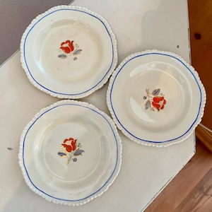 Op de afbeelding: Drie witte keramische borden met blauwe rand en een rood bloemenontwerp. De borden hebben een gegolfde rand.