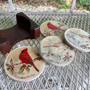 May include: Set of four round coasters with cardinal bird designs and a wooden holder. The coasters feature a red cardinal perched on a branch with holly berries. The holder is a dark brown, curved design.