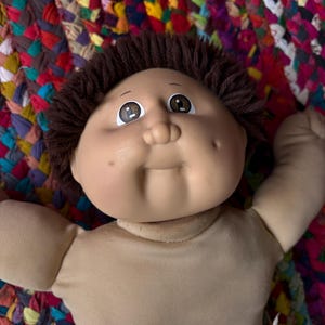 May include: A vintage doll with a tan skin tone, brown hair, and brown eyes. The doll has a soft body and is wearing a tan-colored outfit. The doll is lying on a colorful, woven surface.