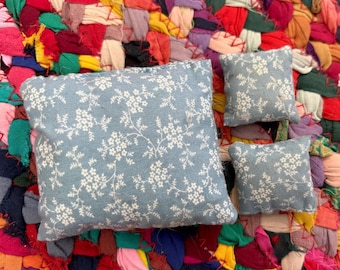 Könnte beinhalten: Drei kleine, quadratische Kissen mit einem hellblauen Blumenmuster. Sie sind auf einem farbenfrohen, strukturierten Teppich aus Stoffresten angeordnet. Das florale Design zeigt zarte weiße Blumen und Ranken.