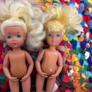 May include: Two vintage dolls with blonde hair and blue eyes. The dolls have a tan skin tone and are posed standing up. The dolls are on a colourful, textured surface.