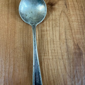 Puede incluir: Una cucharadita de plata con un diseño adornado en el mango. La cuchara tiene un cuenco redondeado y un acabado ligeramente deslustrado.