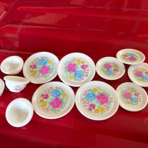 May include: A collection of miniature white dishes, including plates and bowls, arranged on a red surface. The plates feature a floral design with pink, blue, and yellow flowers. The bowls are plain white.
