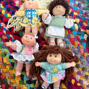 May include: Four vintage Cabbage Patch Kids dolls. Each doll has a unique hairstyle and outfit. One doll wears a blue and yellow plaid dress, another a green and white checkered dress, and two others wear floral dresses. The dolls are on a colorful, woven surface.