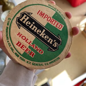May include: A round, brown cardboard coaster with green and black text that reads "IMPORTED Heineken's HOLLAND BEER" and "PRINTED IN BELGIUM". The text is surrounded by a green and white border.