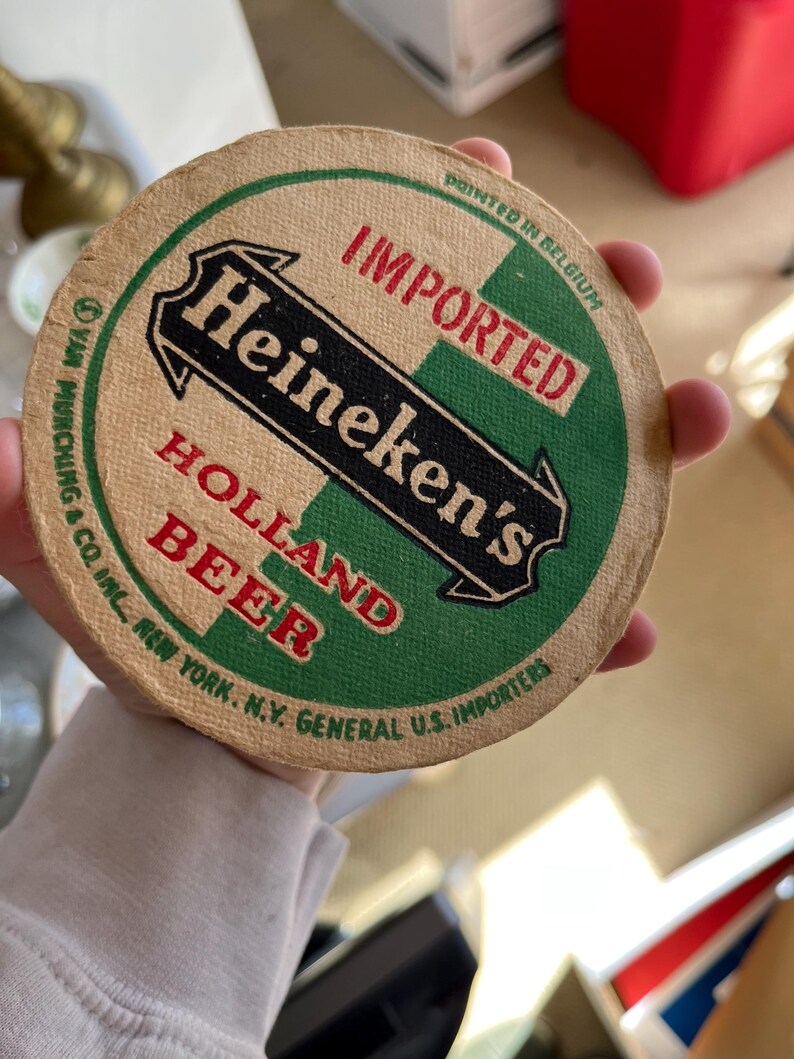 May include: A round, brown cardboard coaster with green and black text that reads "IMPORTED Heineken's HOLLAND BEER" and "PRINTED IN BELGIUM". The text is surrounded by a green and white border.