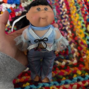 May include: A Cabbage Patch Kids doll with dark hair and brown eyes. The doll is wearing a light blue top with sheer sleeves and denim jeans. The doll's outfit includes a black bow. The doll is a collectible item.