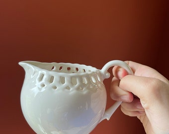 May include: White ceramic creamer with a delicate lace-like design around the top. The creamer has a rounded body and a handle.