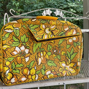 May include: Vintage floral patterned suitcase with a yellow handle. The suitcase features a repeating pattern of large yellow, white, and orange flowers with green leaves on a mustard yellow background. The suitcase has a rectangular shape with a front flap.