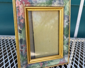 Puede incluir: Un marco de fotos rectangular dorado con un estampado floral en tonos rosa, verde y gris. El marco está hecho de un material dorado y tiene un diseño simple y elegante. El marco está vacío.