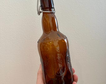 Puede incluir: Una botella de vidrio marrón con una tapa de metal abatible. La botella tiene una superficie texturizada con un diseño de patrón floral.