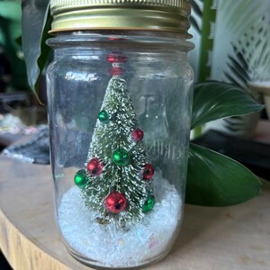 Könnte beinhalten: Ein kleiner Weihnachtsbaum in einem durchsichtigen Glas mit goldenem Deckel. Der Baum ist mit roten und grünen Kugeln geschmückt und steht auf weißem, glitzerndem Schnee. Das Glas steht auf einer Holzoberfläche.