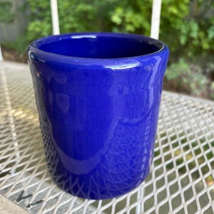 Pode incluir: Um vaso de cerâmica azul cobalto com acabamento brilhante. O vaso cilíndrico tem uma borda ligeiramente alargada e assenta sobre uma superfície de malha metálica branca. O fundo apresenta folhagem verde.