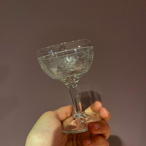 Può includere: Un bicchiere da vino in vetro trasparente con gambo e un motivo inciso decorativo di uva e viti. Il bicchiere è tenuto in una mano.