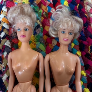 May include: Two vintage fashion dolls with blonde hair and blue eyes. The dolls have a classic design, with articulated arms and legs. The dolls are nude, with a tan skin tone. The dolls are posed side-by-side against a colorful, woven background.