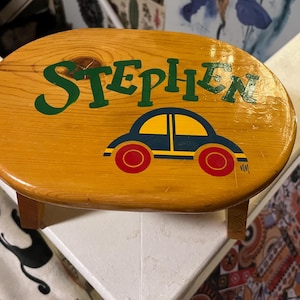 May include: A wooden step stool painted with a blue car and the name "Stephen" in green letters. The stool is oval shaped and has a light wood finish.