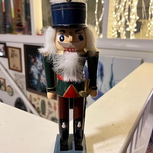 Peut inclure: Une figurine de casse-noisette en bois avec un chapeau bleu, une barbe blanche et un uniforme vert et rouge. Le casse-noisette tient une arme dorée.