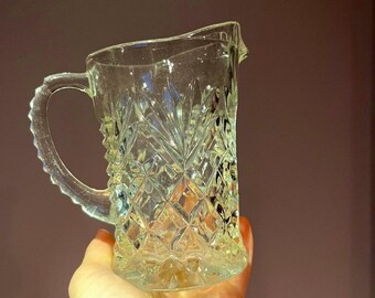 May include: A clear glass pitcher with a cut glass design. The pitcher has a handle and a spout.