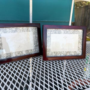 May include: Two rectangular picture frames with a dark reddish-brown finish. Each frame has a light-colored mat with a floral pattern around the opening. The frames are displayed on a white metal surface.