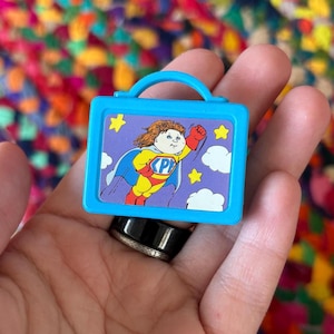 Puede incluir: Una pequeña fiambrera de plástico azul con asa. La fiambrera presenta una imagen de dibujos animados de un superhéroe con capa, volando por el cielo. El superhéroe tiene una "P" en el pecho y está rodeado de estrellas y nubes.