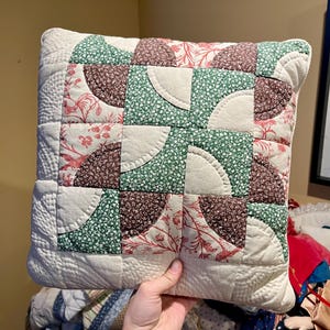 May include: A white pillow with a quilted design featuring a patchwork of green and brown fabric with a floral pattern. The pillow is made of cotton and has a square shape.