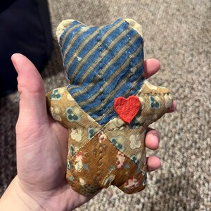 May include: A handmade fabric bear with a blue and gold striped head and a red heart. The bear's body is a patchwork of floral and patterned fabrics in shades of brown and beige. A charming, handcrafted item.