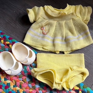 Könnte beinhalten: Gelbes Strick-Baby-Outfit mit Enten-Applikation, weißen Streifen und passenden Bloomers. Das Set enthält ein Paar weiße Mary Jane Schuhe. Das Outfit ist für ein Baby.