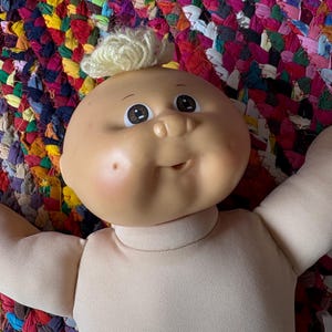 May include: A vintage Cabbage Patch Kid doll with a round, peach-colored face, rosy cheeks, and a small tuft of blonde hair. The doll has large, expressive eyes and a smiling mouth. The doll's body is a light beige color.