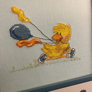 K&ouml;nnte beinhalten: Eine gerahmte Kreuzstich-Stickerei mit einem gelben K&uuml;ken, das zwei Ballons h&auml;lt. Ein Ballon ist blau und der andere orange. Das K&uuml;ken tr&auml;gt blaue Schuhe und steht auf gr&uuml;nem Gras.