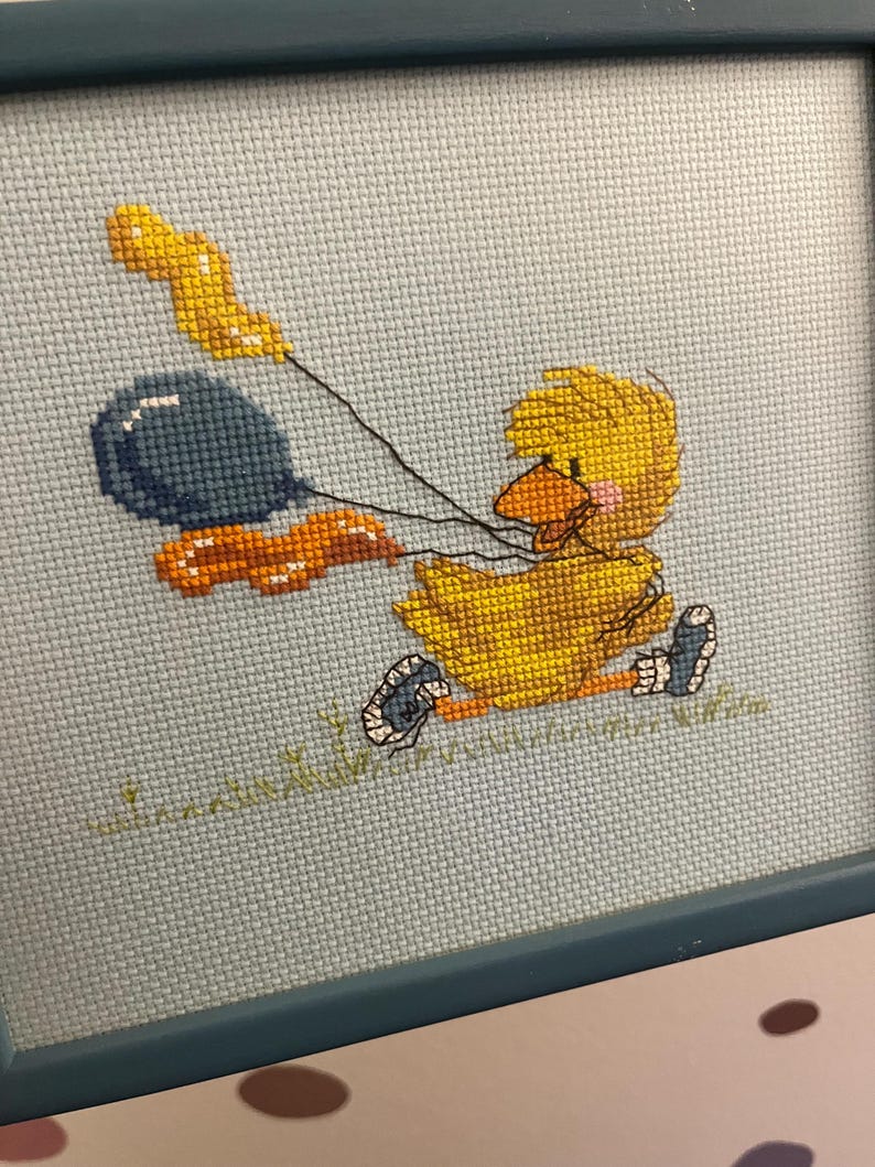 K&ouml;nnte beinhalten: Eine gerahmte Kreuzstich-Stickerei mit einem gelben K&uuml;ken, das zwei Ballons h&auml;lt. Ein Ballon ist blau und der andere orange. Das K&uuml;ken tr&auml;gt blaue Schuhe und steht auf gr&uuml;nem Gras.
