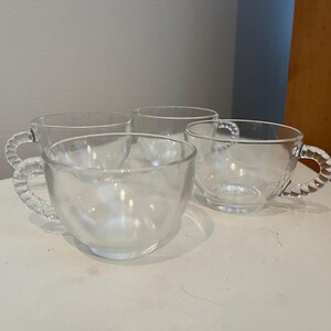 Puede incluir: Cuatro tazas de vidrio transparente con asas retorcidas. Las tazas están vacías y están sobre una superficie blanca.