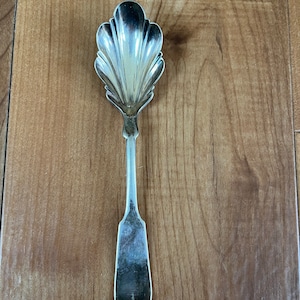 May include: A silver sugar spoon with a scalloped bowl and a simple, rounded handle.