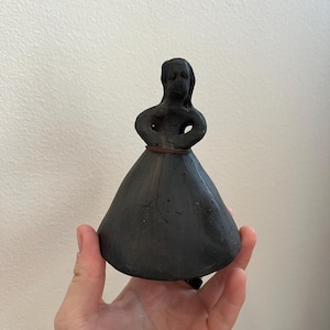 May include: Black ceramic figurine of a woman wearing a long dress. The figurine is approximately 15 cm tall and has a simple, rustic design.