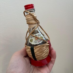 May include: A small glass bottle of Italian Swiss Colony California White Wine, wrapped in a woven straw basket with a red base. The bottle has a red cap and a label with the brand name and a Swiss flag.