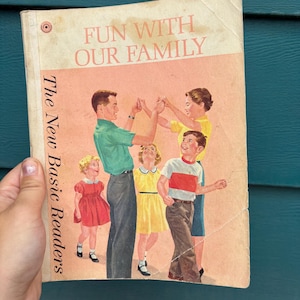 May include: Vintage book cover titled "Fun With Our Family" featuring an illustration of a family dancing. The book is part of "The New Basic Readers" series. The cover has a light pink background with the family illustration.