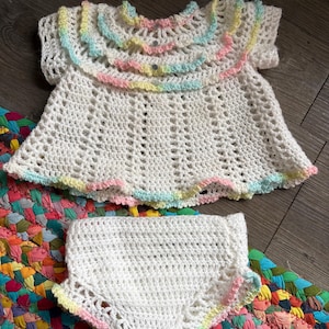 May include: A white crochet baby dress and matching diaper cover set. The dress features a ruffled collar and sleeves, with pastel rainbow trim. The diaper cover has matching trim. The set is displayed on a dark wood surface.