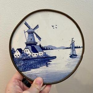 May include: A round ceramic plate with a blue and white painted scene of a windmill, houses, and a sailboat on a body of water. The plate has a brass rim.