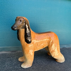 Puede incluir: Figura de cerámica de un galgo afgano en tonos marrón, negro y beige. El perro tiene pelo largo y está de pie sobre cuatro patas. Es un objeto decorativo.