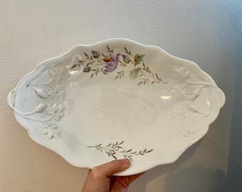 May include: White oval serving dish with a floral design in shades of purple, green, and brown. The dish has a raised rim and a scalloped edge.
