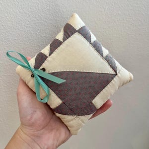 May include: A small, square, quilted pillow with a light brown and dark gray patchwork design. The pillow has a light blue ribbon tied around it.