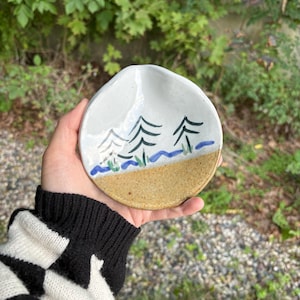 May include: Handmade ceramic dish with a landscape design. The dish features a white upper section with painted green trees and blue water, transitioning to a tan lower section. The dish is round with a slightly irregular shape.