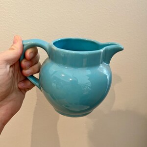 Puede incluir: Una jarra de cerámica azul claro con cuerpo redondeado y asa. La jarra está hecha de cerámica esmaltada y tiene un acabado liso y brillante.