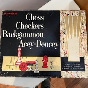 Puede incluir: Una caja de juego vintage con fondo negro y texto blanco. La caja presenta el título "Chess Checkers Backgammon Acey-Deucey". La caja también incluye imágenes de piezas de juego, incluyendo un caballo de ajedrez rojo, un peón de ajedrez blanco y una ficha de damas roja.