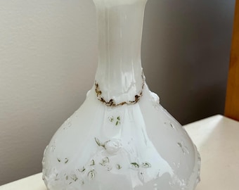Puede incluir: Un jarrón de vidrio blanco con un delicado diseño floral y detalles dorados. El jarrón tiene un cuerpo redondo y un cuello estrecho.