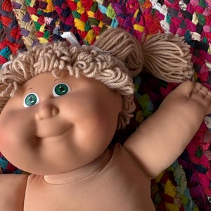 May include: A vintage Cabbage Patch Kid doll with tan skin, green eyes, and light brown yarn hair styled in pigtails. The doll is wearing a tan fabric body. The background is a colorful, woven rug.