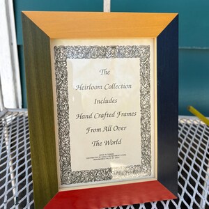 May include: A multi-coloured picture frame with a rectangular opening. The frame is made of wood and features a combination of colours, including yellow, green, red, and navy blue. The frame contains text that reads "The Heirloom Collection Includes Hand Crafted Frames From All Over The World."