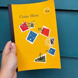 May include: A vintage yellow notebook with the words "Come Here" printed on the cover. The cover features colorful geometric shapes and illustrations of a fishbowl, helicopter, cat, stars, sun, sailboat, and clouds. The notebook has a black spine.